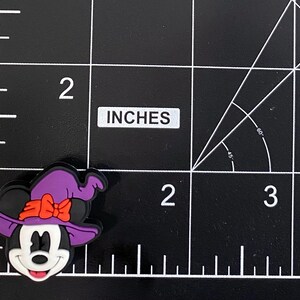 May include: A colourful charm featuring a cartoon mouse with a witch hat and bow. The charm is black, white, purple, and orange. The charm is approximately 5 cm in size.