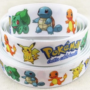 Op de afbeelding: Witte lint met kleurrijke stripfiguren en het woord "Pokémon" in blauw en geel. Het lint toont afbeeldingen van Bulbasaur, Squirtle, Charmander en Pikachu. De tekst "Gotta catch 'em all!" is ook aanwezig.