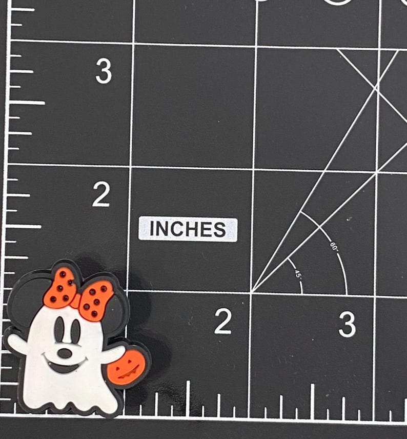 K&ouml;nnte beinhalten: Ein Halloween-Motiv-Charm mit einem Cartoon-Geist mit Minnie-Maus-Ohren und einer Schleife. Der Geist ist wei&szlig; mit einem l&auml;chelnden Gesicht und h&auml;lt einen orangefarbenen K&uuml;rbis. Der Charm ist etwa 2,5 cm gro&szlig;.