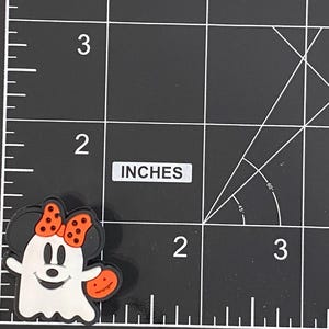 May include: A Halloween-themed charm featuring a cartoon ghost with Minnie Mouse ears and a bow. The ghost is white with a smiling face and is holding an orange pumpkin. The charm is approximately 2.5 cm in size.