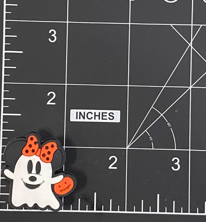 May include: A Halloween-themed charm featuring a cartoon ghost with Minnie Mouse ears and a bow. The ghost is white with a smiling face and is holding an orange pumpkin. The charm is approximately 2.5 cm in size.