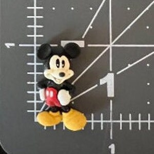 Puede incluir: Peque&ntilde;a figura tridimensional de Mickey Mouse. La figura tiene orejas negras, cuerpo negro, pantalones cortos rojos, zapatos amarillos y guantes blancos. La figura est&aacute; sobre una superficie gris con marcas de medici&oacute;n blancas.
