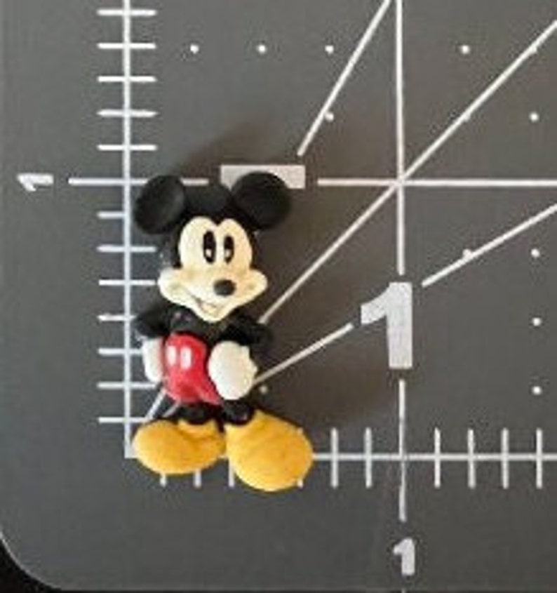 Puede incluir: Peque&ntilde;a figura tridimensional de Mickey Mouse. La figura tiene orejas negras, cuerpo negro, pantalones cortos rojos, zapatos amarillos y guantes blancos. La figura est&aacute; sobre una superficie gris con marcas de medici&oacute;n blancas.