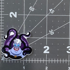K&ouml;nnte beinhalten: Ein lila und schwarzer Ursula-Aufkleber mit wei&szlig;em Hintergrund. Ursula ist eine Figur aus dem Disney-Film Die kleine Meerjungfrau. Sie ist eine Meereshexe mit langen wei&szlig;en Haaren, einem lila Kleid und einer Halskette mit einem goldenen Anh&auml;nger. Sie hat ein b&ouml;ses L&auml;cheln und ist von Tentakeln umgeben.