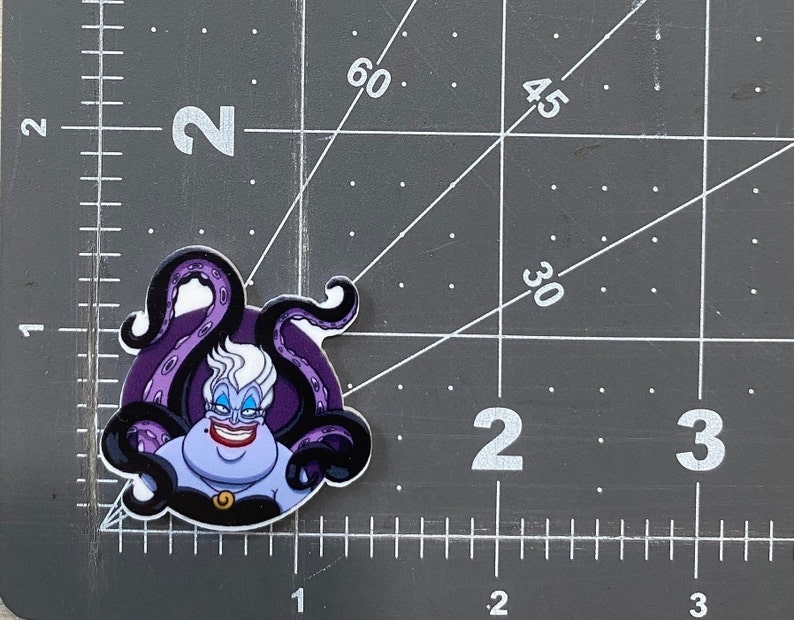K&ouml;nnte beinhalten: Ein lila und schwarzer Ursula-Aufkleber mit wei&szlig;em Hintergrund. Ursula ist eine Figur aus dem Disney-Film Die kleine Meerjungfrau. Sie ist eine Meereshexe mit langen wei&szlig;en Haaren, einem lila Kleid und einer Halskette mit einem goldenen Anh&auml;nger. Sie hat ein b&ouml;ses L&auml;cheln und ist von Tentakeln umgeben.