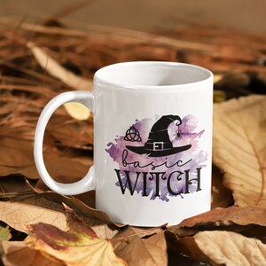 May include: White ceramic mug with a purple and pink watercolor design featuring a black witch hat with a spider and the text "basic witch".