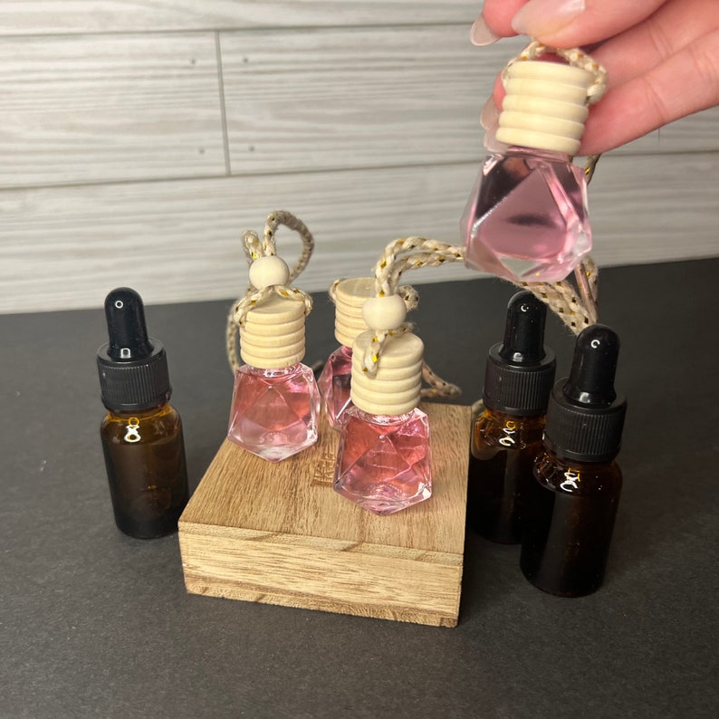 May include: A set of four pink glass car air fresheners with wooden tops and gold string. The fresheners are on a wooden stand. There are three additional brown glass bottles with droppers.