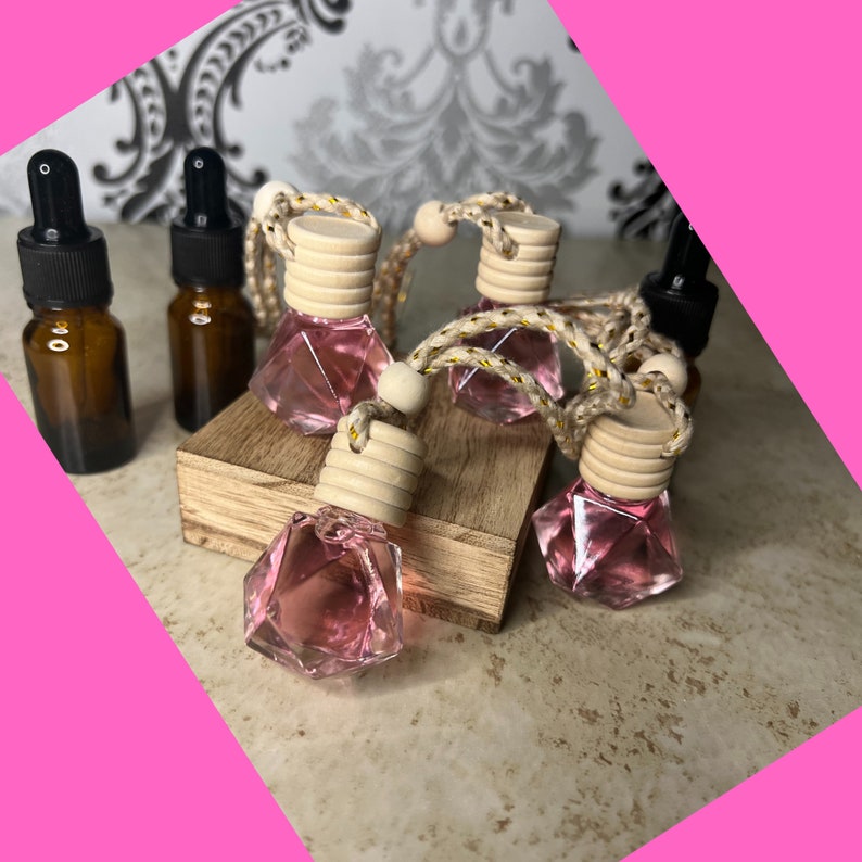 May include: Five pink glass car air fresheners with wooden caps and gold string hanging from them. The air fresheners are shaped like diamonds and are sitting on a wooden surface.