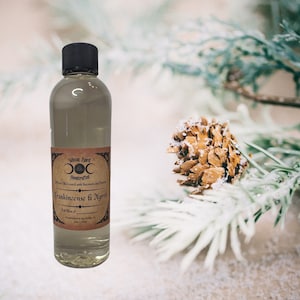 May include: A clear glass bottle with a black cap and a label that reads "Frankincense & Myrrh" with a triple moon symbol and the text "Vircan Spirit Handcrafted" in a vintage style font. The bottle is sitting on a white surface with a pine cone and pine needles in the background.