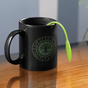 May include: Black ceramic mug with a green tree of life design surrounded by runes. The mug has a black handle and is filled with a dark liquid. A green plant is sticking out of the mug.