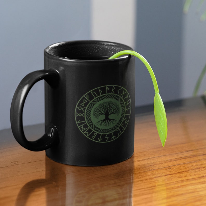 May include: Black ceramic mug with a green tree of life design surrounded by runes. The mug has a black handle and is filled with a dark liquid. A green plant is sticking out of the mug.