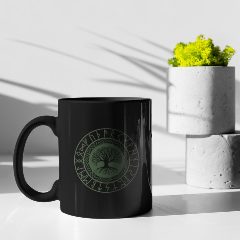May include: Black ceramic mug with a green tree of life design surrounded by a circle of runes. The mug is on a white surface.