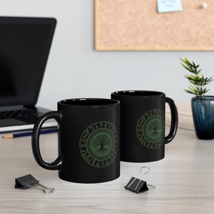 May include: Two black ceramic mugs with a green tree of life design and runes around the tree. The mugs are on a wooden surface.