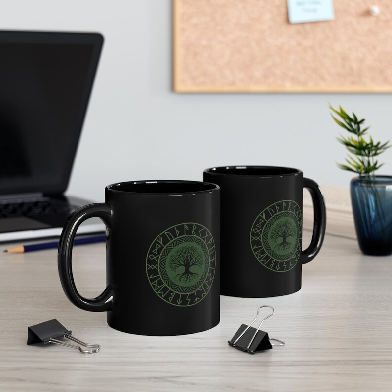 May include: Two black ceramic mugs with a green tree of life design and runes around the tree. The mugs are on a wooden surface.
