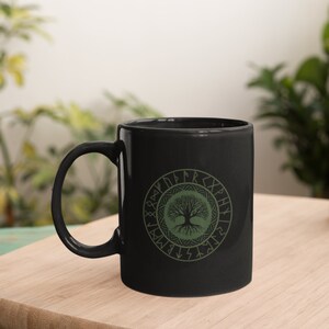 May include: Black ceramic mug with a green tree of life design surrounded by a circle of runes. The runes are in a light green color.