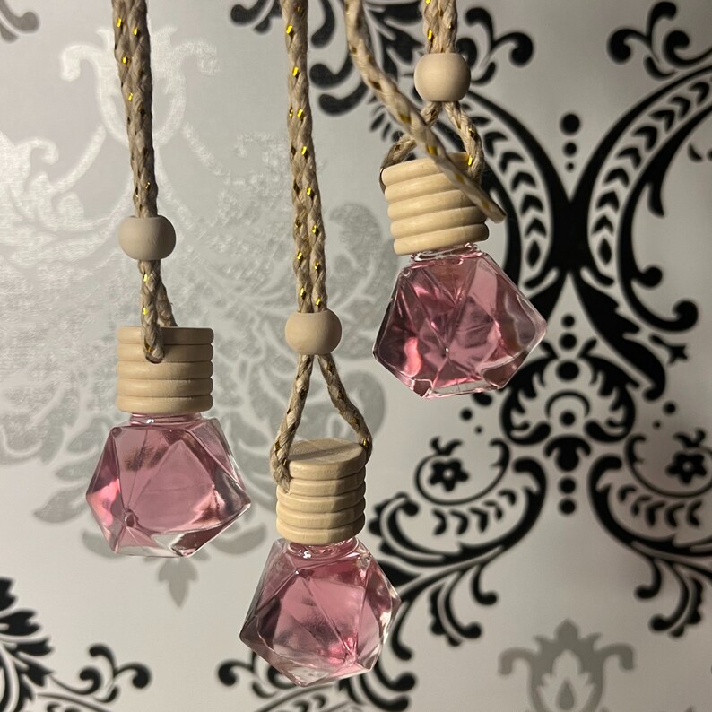 May include: Three pink glass car air fresheners with wooden caps hanging from brown cords. The fresheners are shaped like diamonds and are filled with a pink liquid.