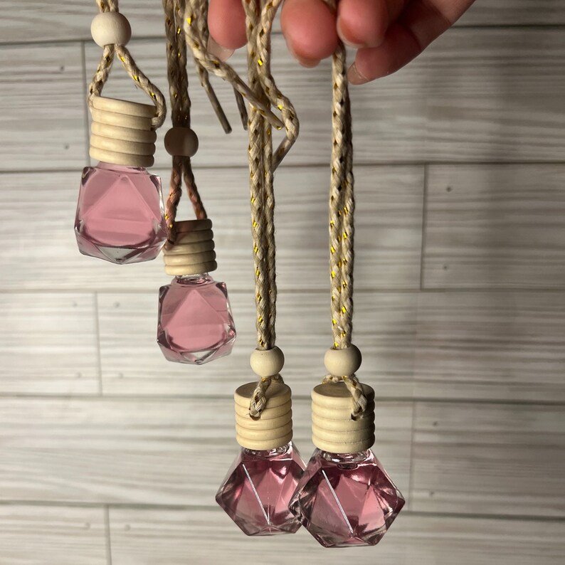 May include: Four pink glass car air fresheners with wooden tops and gold string hangers. The fresheners are shaped like a diamond and filled with a pink liquid.