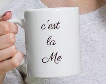 May include: White ceramic mug with black text that reads "c'est la Me".