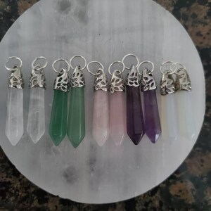 May include: Eight crystal point pendants with silver caps, displayed on a white circular surface. The pendants are in various colors, including clear, white, green, pink, purple, and light yellow.