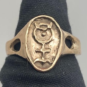 May include: A gold-toned ring with an oval-shaped design featuring a stylized symbol with a cross and wings. The ring has two cut-out holes on the sides.