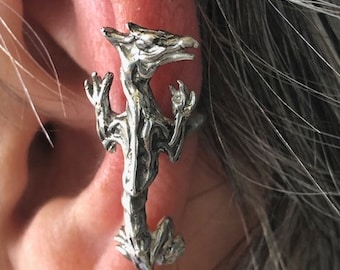 May include: A silver-toned dragon ear cuff. The dragon is detailed with textured scales and a stylized form, appearing to wrap around the ear. The cuff is designed to be worn on the ear, adding a unique accessory.