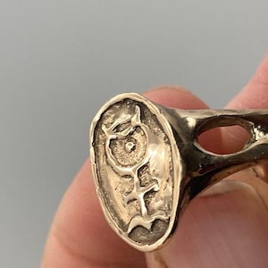 May include: A gold ring with a unique design featuring a stylized symbol on the face of the ring. The ring has a wide band with an open design.