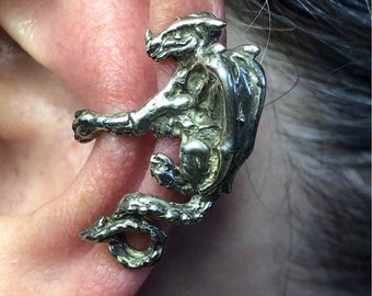 May include: Silver dragon ear cuff. The dragon is curled up with its wings spread out. It is a detailed piece of jewelry.