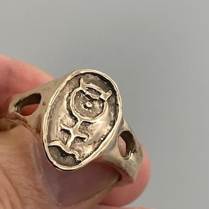 May include: A gold-toned ring with an oval-shaped design featuring a stylized symbol. The ring has two cut-out holes on either side of the oval.