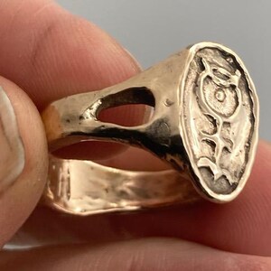 May include: A gold ring with a raised oval design featuring a stylized fish with a circle in the center. The ring has a wide band with a cutout.