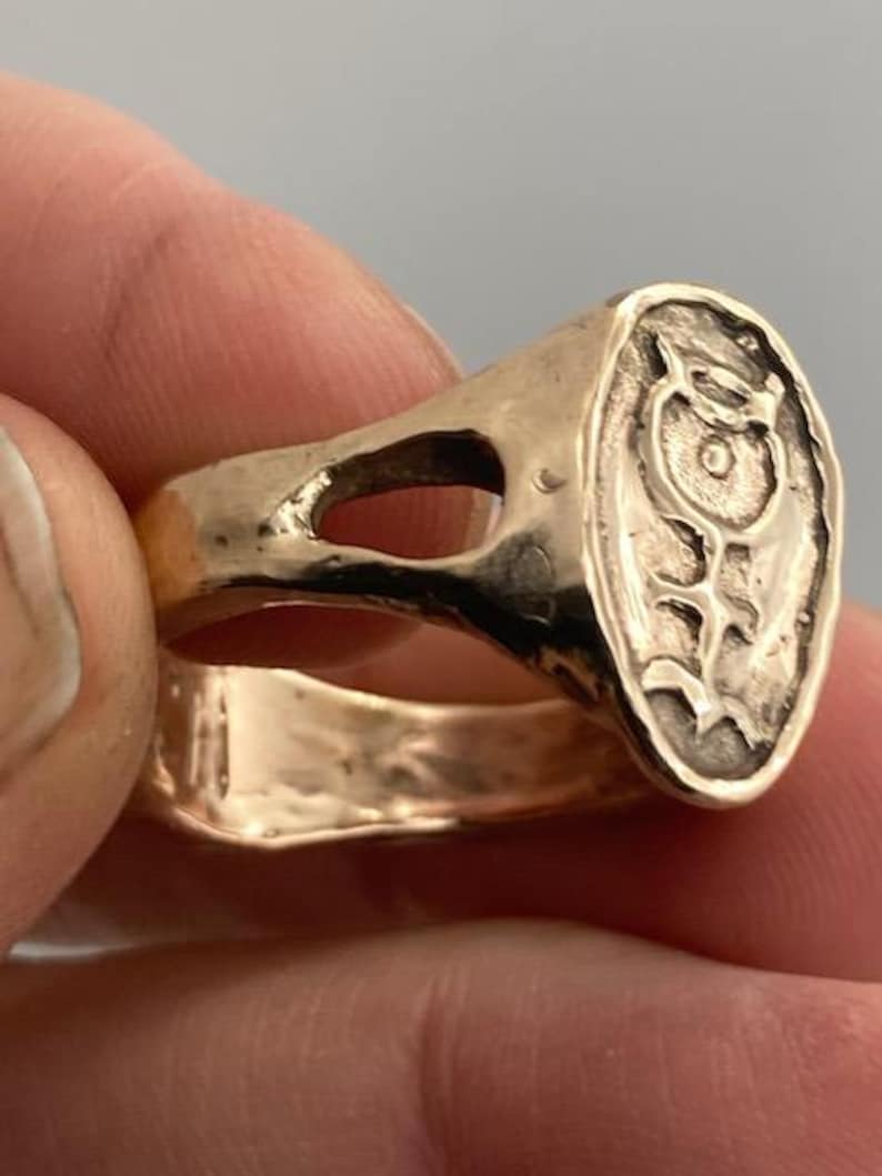 May include: A gold ring with a raised oval design featuring a stylized fish with a circle in the center. The ring has a wide band with a cutout.
