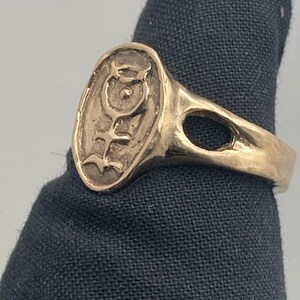 May include: A gold ring with an oval-shaped design featuring a stylized symbol with a circle and lines. The ring has a smooth, polished finish.
