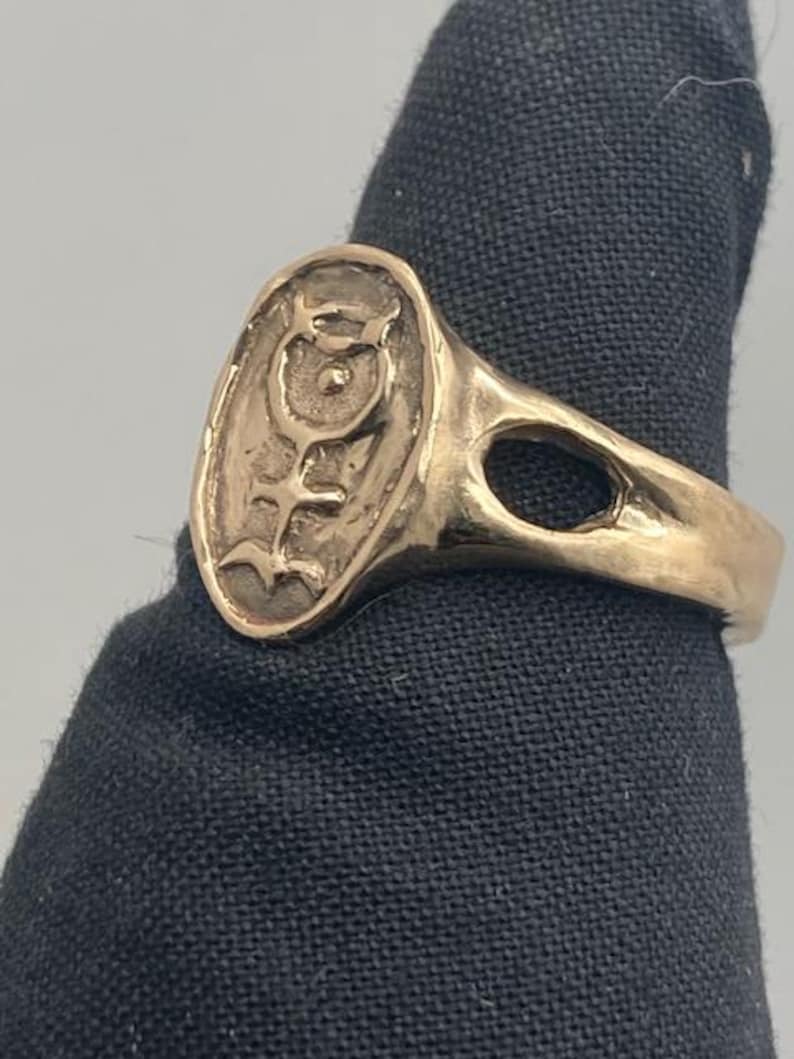 May include: A gold ring with an oval-shaped design featuring a stylized symbol with a circle and lines. The ring has a smooth, polished finish.