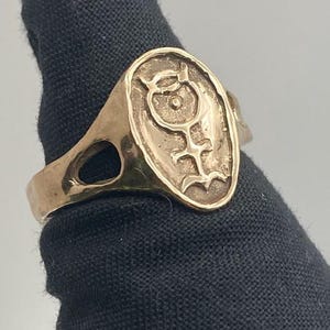 May include: A gold ring with an oval-shaped design featuring a stylized symbol. The symbol is a circle with a dot in the center and a line extending from the bottom.
