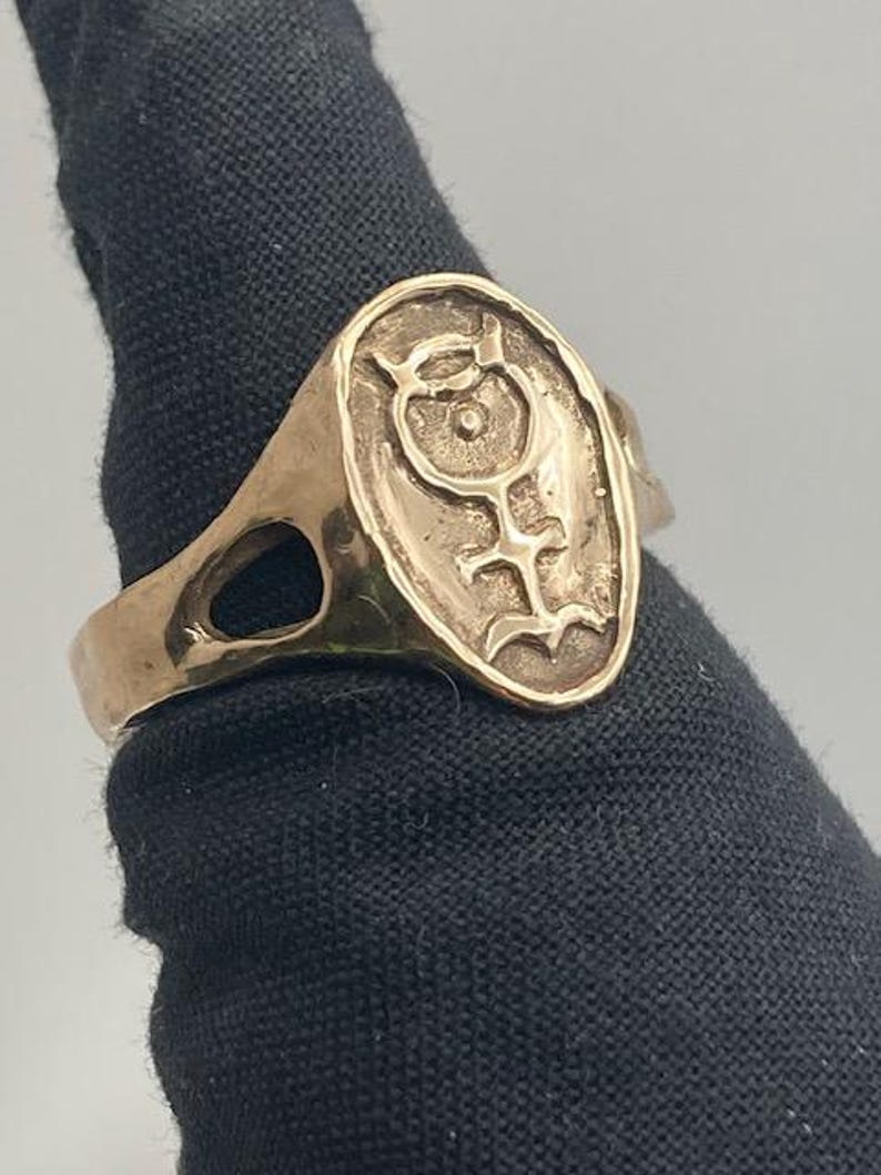May include: A gold ring with an oval-shaped design featuring a stylized symbol. The symbol is a circle with a dot in the center and a line extending from the bottom.