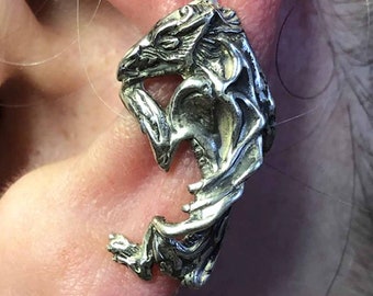 May include: A silver-toned earring in the shape of a mythical creature, possibly a dragon or gargoyle. The earring is detailed with intricate textures and features a stylized design. The earring is worn on an ear.