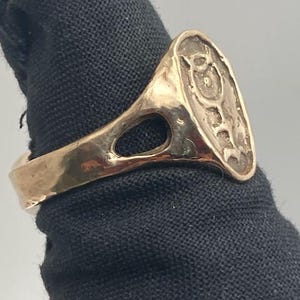 May include: A gold-colored ring with a large oval-shaped top. The top of the ring has an intricate design with a raised center.