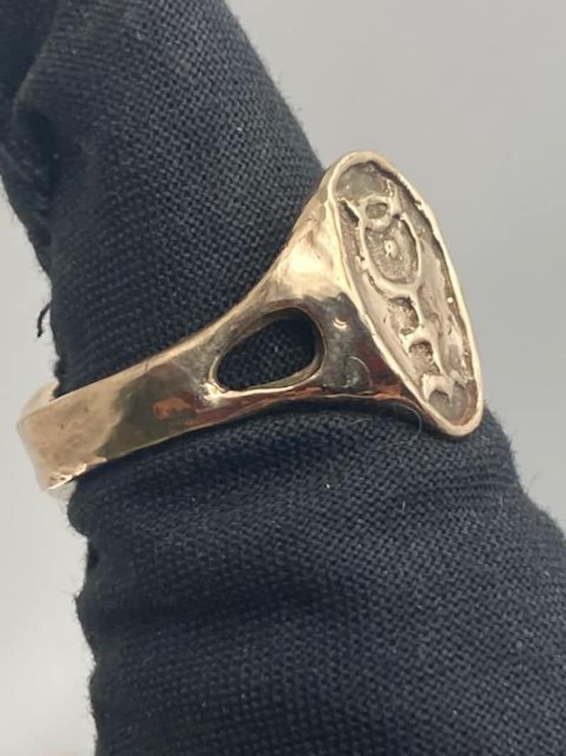May include: A gold-colored ring with a large oval-shaped top. The top of the ring has an intricate design with a raised center.