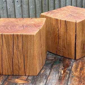 May include: Two square wooden stools crafted from solid logs, featuring a natural light brown finish with visible wood grain patterns, distinct growth rings on the top surfaces, and deep vertical cracks running along the sides of each block.