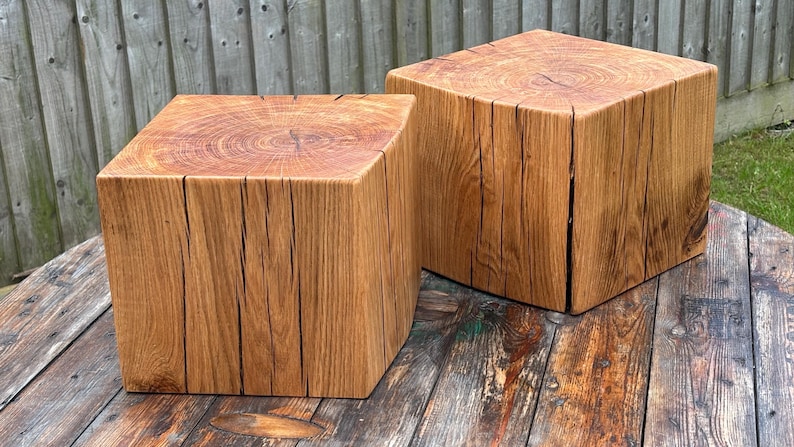 May include: Two square wooden stools crafted from solid logs, featuring a natural light brown finish with visible wood grain patterns, distinct growth rings on the top surfaces, and deep vertical cracks running along the sides of each block.