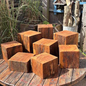 May include: A collection of seven solid wooden blocks with a natural, reddish-brown finish. Each block features a distinct cubic shape, visible wood grain patterns, and deep vertical cracks along the sides, showcasing the raw texture of the timber.