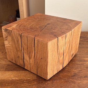 May include: A square-shaped block of natural wood with a visible grain pattern on the top surface. The sides feature vertical wood grain textures and several deep, natural cracks running from the top edge down toward the base of the block.