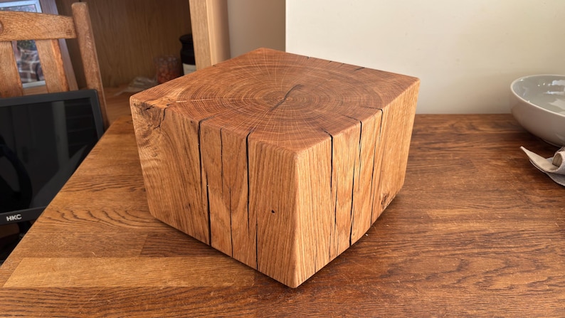May include: A square-shaped block of natural wood with a visible grain pattern on the top surface. The sides feature vertical wood grain textures and several deep, natural cracks running from the top edge down toward the base of the block.