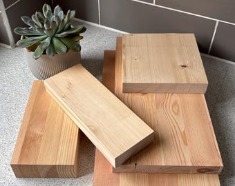 May include: A collection of unfinished, light-colored wooden blocks and planks. The wood grain is visible, and the pieces vary in size and shape. A potted succulent plant with green leaves sits nearby. The blocks are arranged on a speckled surface.
