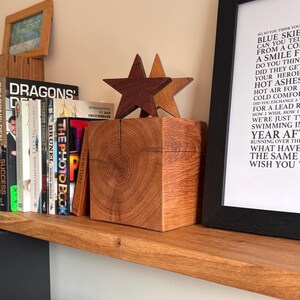 May include: A square wooden block with a visible grain pattern, topped with two five-pointed stars in dark and light brown wood. The block sits on a wooden shelf next to a row of books and a framed print featuring lyrics from the song Wish You Were Here.