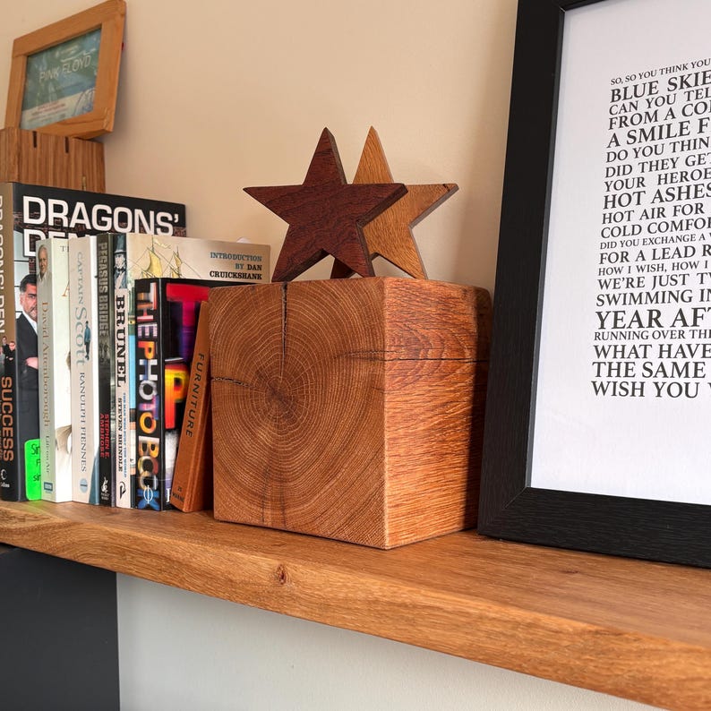 May include: A square wooden block with a visible grain pattern, topped with two five-pointed stars in dark and light brown wood. The block sits on a wooden shelf next to a row of books and a framed print featuring lyrics from the song Wish You Were Here.