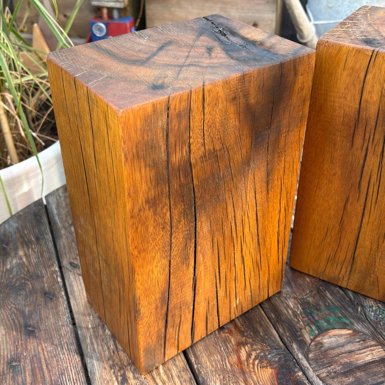 May include: Two rectangular blocks of natural wood with a warm amber-brown tone. The surfaces display prominent vertical grain patterns, deep dark brown fissures, and a rough, weathered texture across the sides and the flat top faces.
