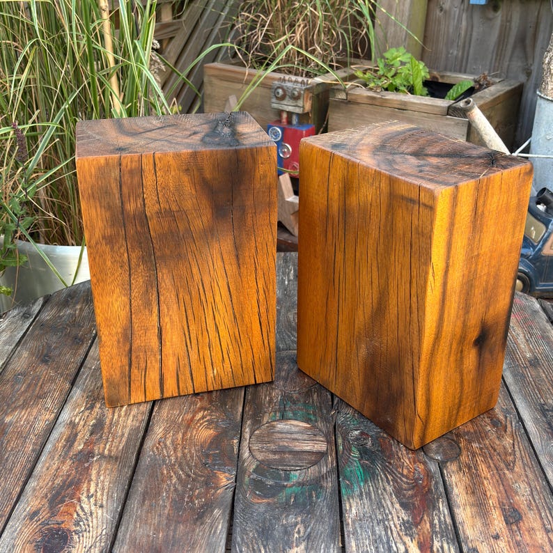 May include: Two rectangular blocks of natural wood with a warm brown finish, featuring visible vertical grain patterns, deep cracks, and dark knots. The blocks have a smooth, polished surface and are positioned upright, showcasing the raw texture of the timber.