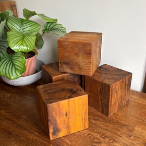 May include: A collection of four solid wooden cubes with a natural brown finish and visible wood grain patterns. The cubes are arranged in a stacked formation, showcasing the smooth, square-cut edges and the rich, warm tones of the timber material.