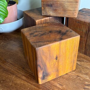 May include: A collection of four solid wooden cubes with a natural brown finish. Each cube displays a distinct wood grain pattern, visible knots, and fine surface cracks. The blocks are arranged in a cluster, showcasing the varied textures and warm, earthy tones of the wood material in a square, geometric shape.