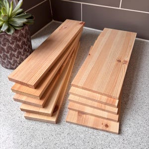 May include: Two stacks of unfinished wooden planks. The planks are a light brown colour with visible wood grain. The rectangular planks are ready for a woodworking project. Each plank is approximately 2.5 cm thick.